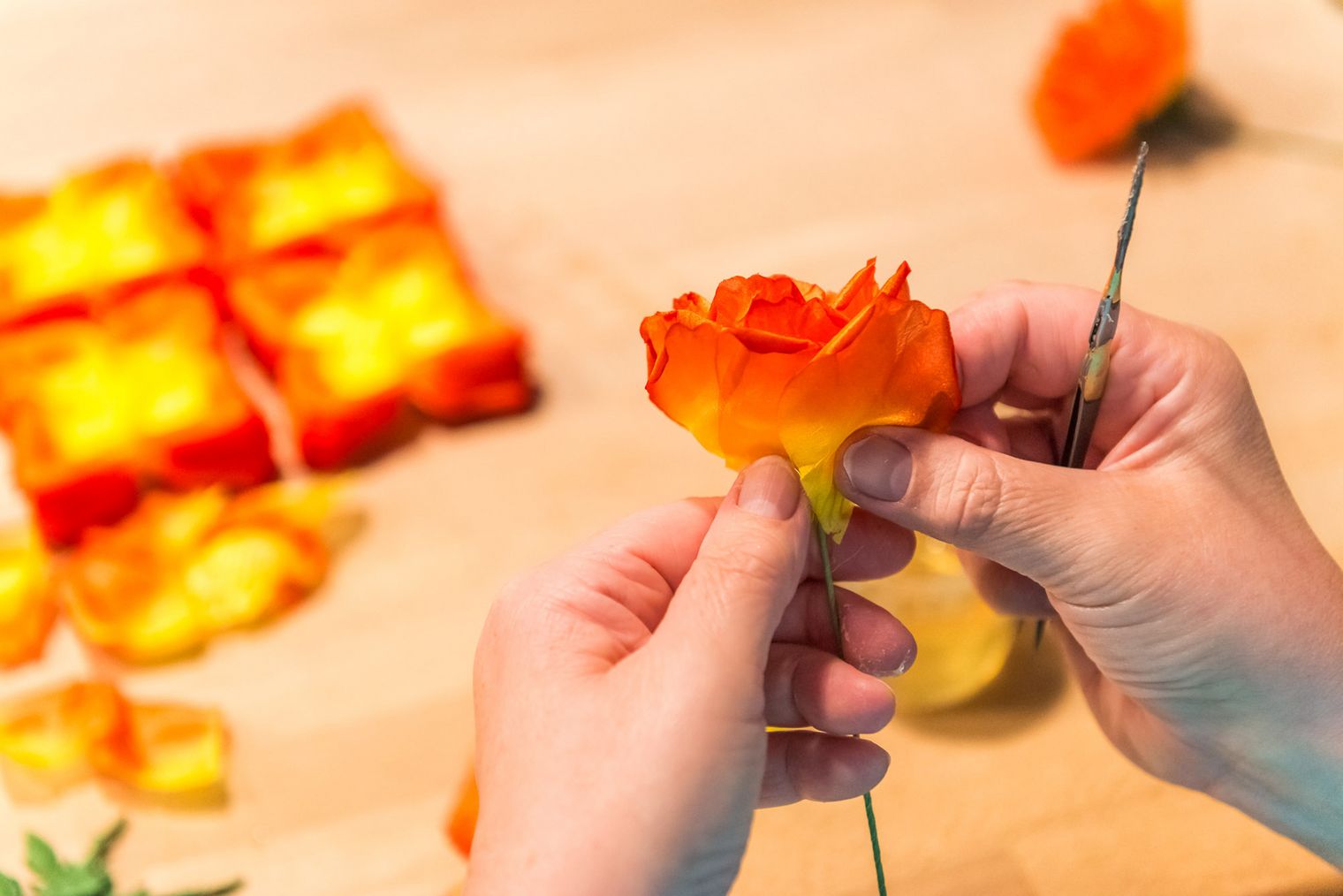 Hände formen eine orange-blühende Blume aus buntem Material, umgeben von weiteren Blüten und quadratischen Formen.