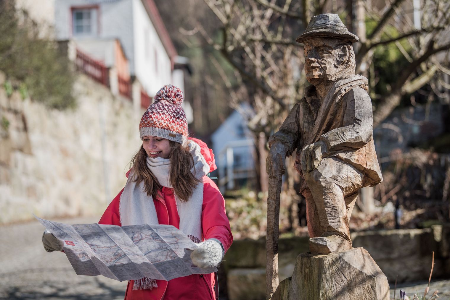 Frau in rotem Mantel und Schal hält eine Wanderkarte vor einer Holzskulptur eines Mannes.