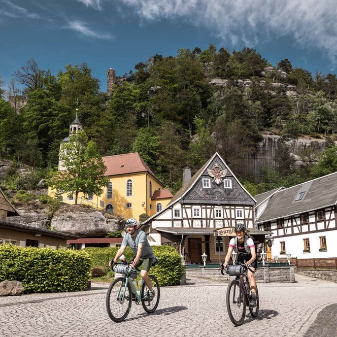 Zwei Radfahrer fahren auf einem gepflasterten Weg vor traditionellen Gebäuden und einem bewaldeten Hügel.