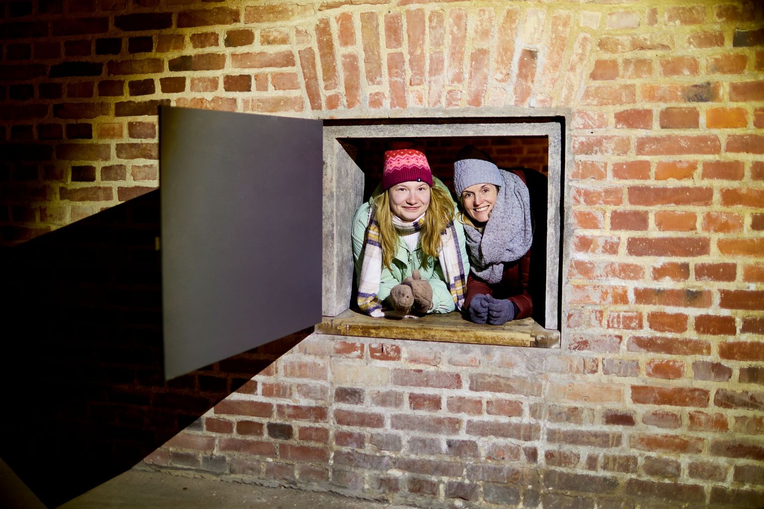 Zwei Personen schauen neugierig aus einem kleinen Fenster in einer Ziegelmauer.