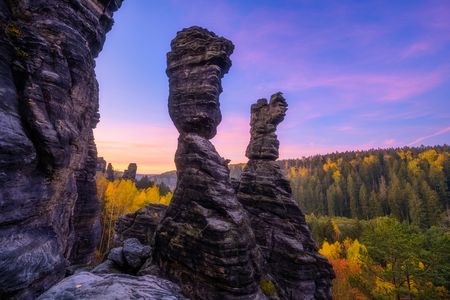 Felsformationen mit markanten, säulenartigen Strukturen vor einem bunten Sonnenuntergang und bewaldeter Landschaft.