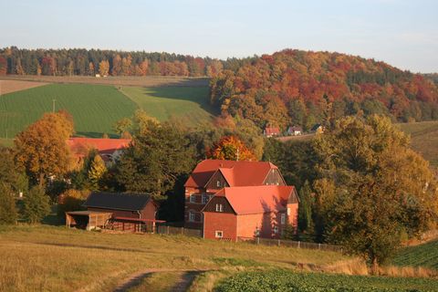 Landschaft mit einem roten Bauernhaus, umgeben von Bäumen mit buntem Herbstlaub und sanften Hügeln.