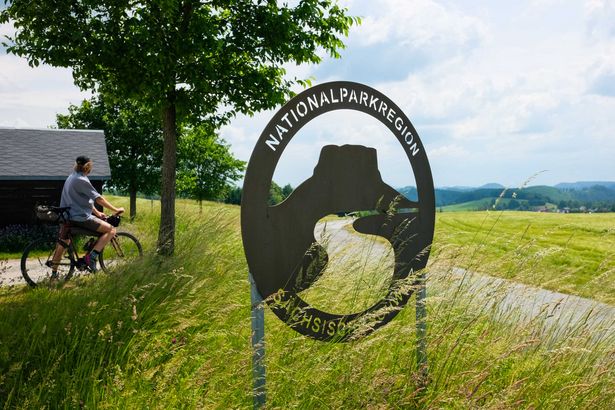 Ein gruenes Schild und die Aufschrift "Nationalparkregion Sächsische Schweiz", im Hintergrund gruene Wiesen.