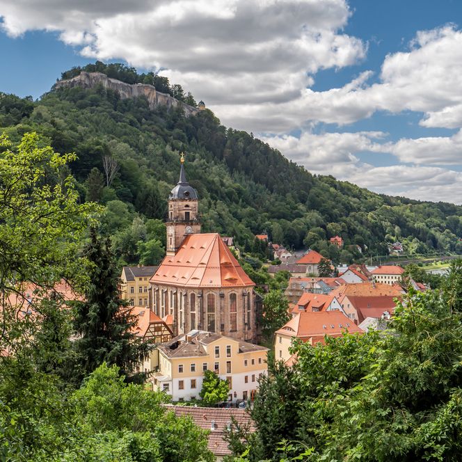 Blick auf eine Stadt mit einer Kirche und der Festung Königstein im Hintergrund, umgeben von Bäumen und Wolken.
