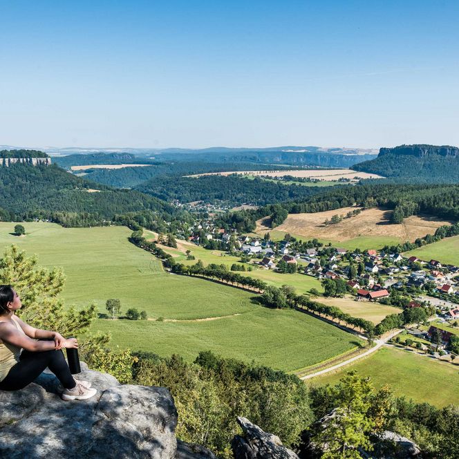 Person sitzt auf einem Felsen und blickt auf eine grüne Landschaft mit Dörfern und Hügeln.