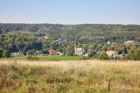 Landschaft mit Feldern, Bäumen und einem kleinen Dorf im Hintergrund unter blauem Himmel.
