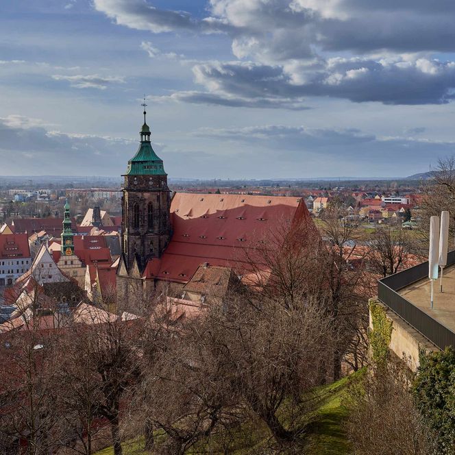 Blick auf eine Stadt mit roten Dächern, einer Kirche mit grünem Turm und bewölktem Himmel.