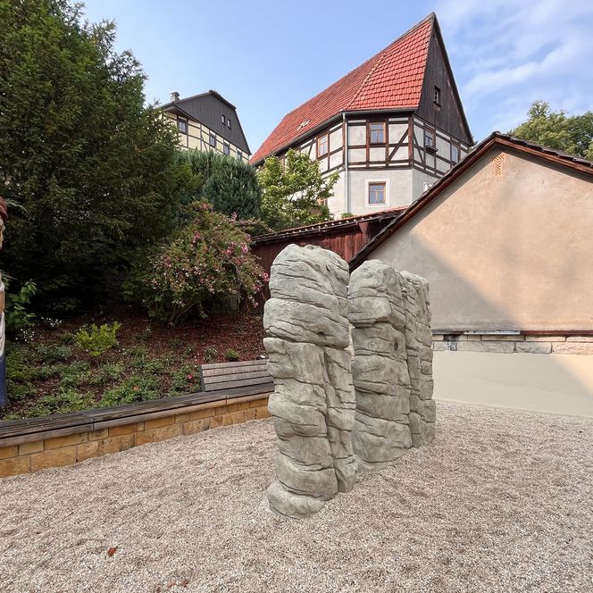 Zwei kleine Kletterfelsen stehen in einem kleinen Park, im Hintergrund ein Fachwerkhaus und ein totemähnlicher Holzschnitt.