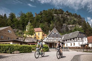 Zwei Radfahrer fahren auf einem gepflasterten Weg vor traditionellen Gebäuden und einem bewaldeten Hügel.
