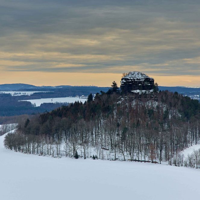 Schneebedeckter Hügel mit Bäumen unter einem bewölkten Himmel bei Dämmerung.