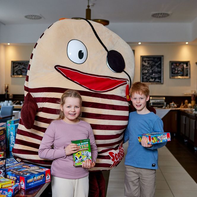 Zwei Kinder stehen neben einem großen, gestreiften Maskottchen mit Augenklappe und breitem Lächeln.