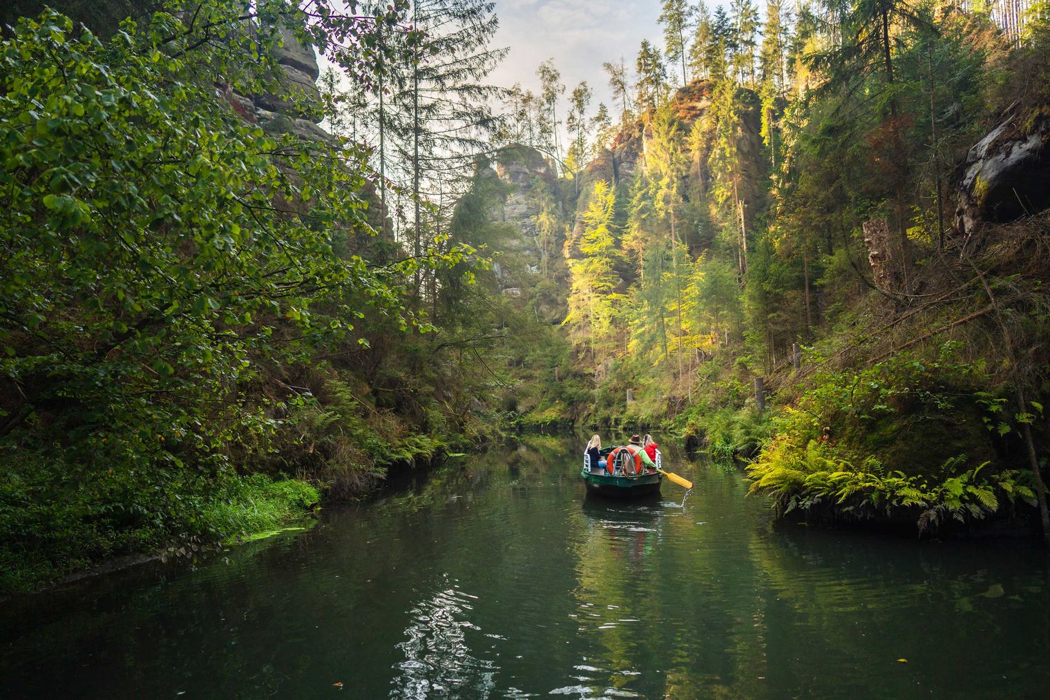 Ruderboot auf einem ruhigen Fluss, umgeben von grünen Bäumen und steilen Felsen.