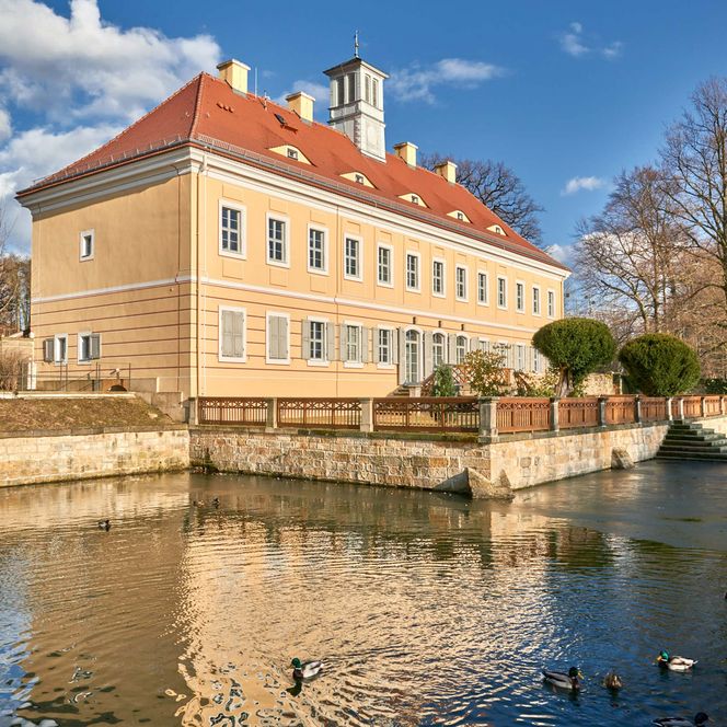 Historisches Gebäude mit rotem Dach, umgeben von Wasser und Bäumen, unter blauem Himmel mit Wolken.
